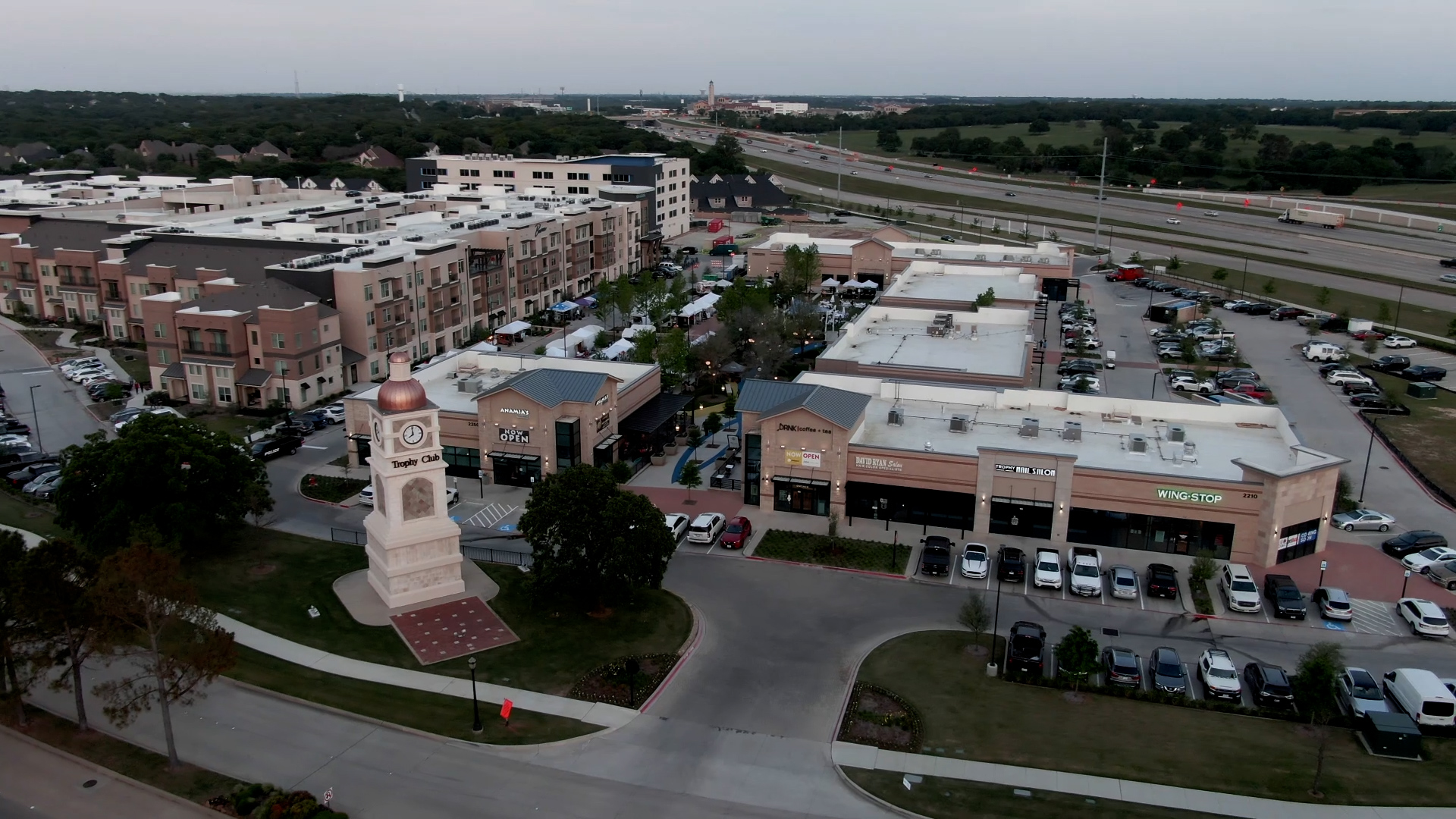 Aerial view of Trophy Club Town Center at dusk, with the Trophy Club clock tower, shops, restaurants, and Highway 114 in the distance