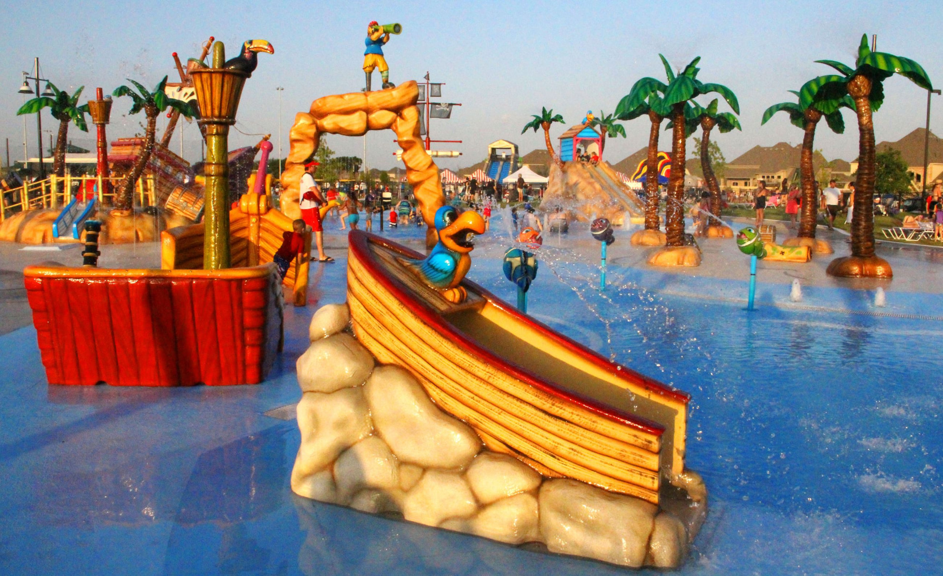 Children playing at the splash pad among boat slides, water sprayers, and palm tree features on a busy summer day