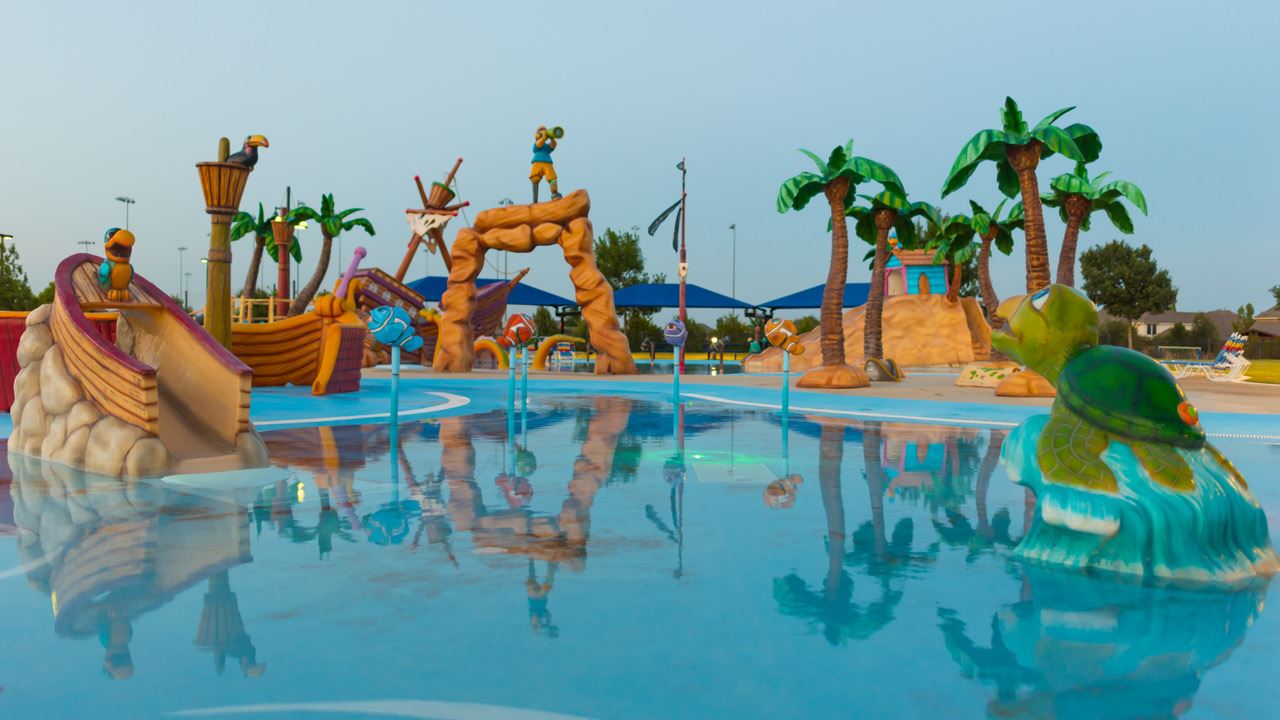A pirate-themed splash pad with palm tree sprayers, slides, and shallow water reflecting the evening sky