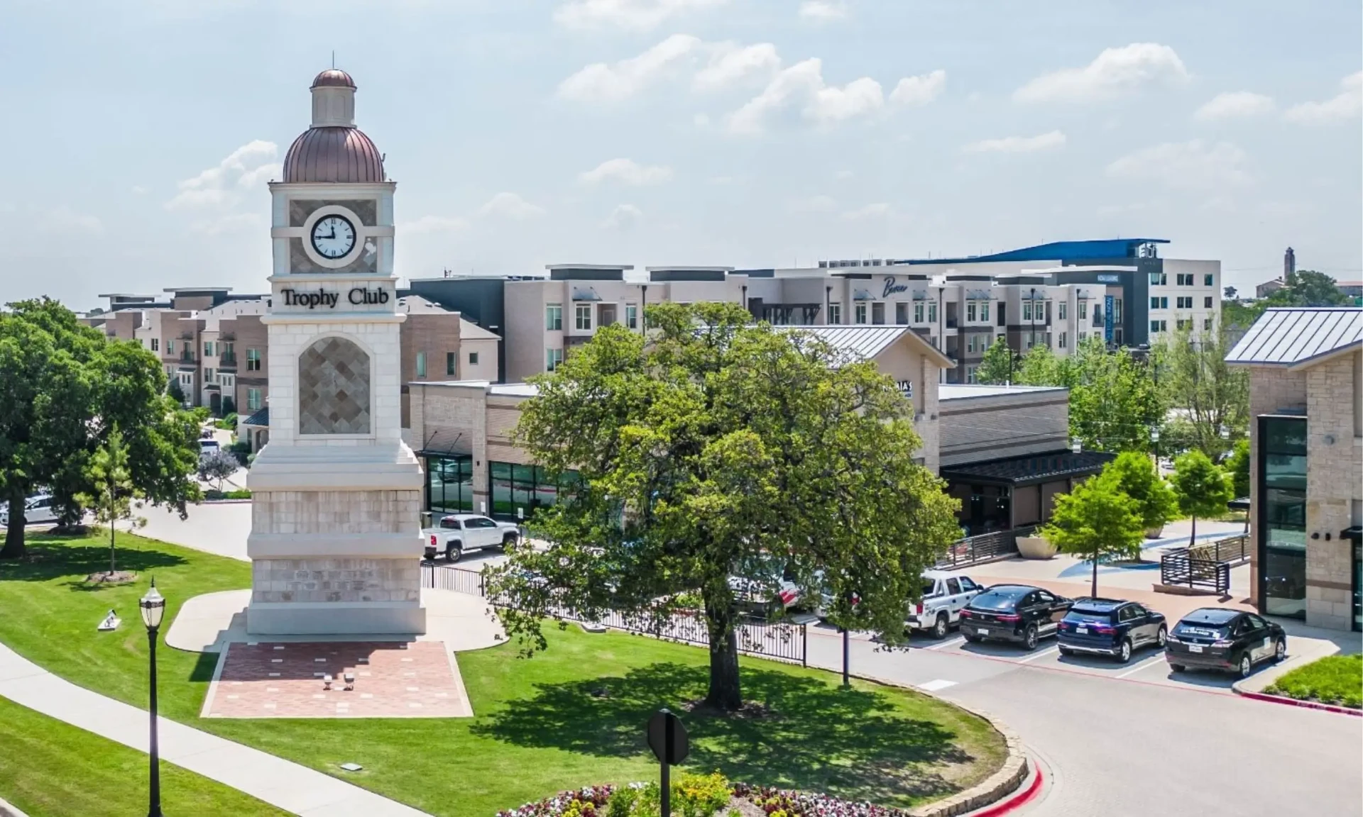 The Trophy Club clock tower beside a large oak tree, with Town Center shops, restaurants, and apartments behind it
