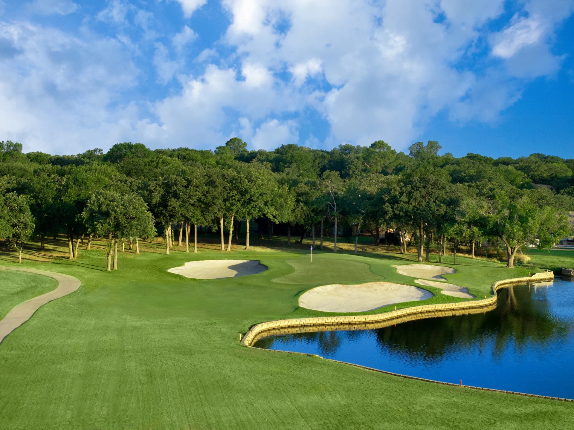 A manicured golf green framed by bunkers, mature oak trees, and a pond on a sunny day in Trophy Club