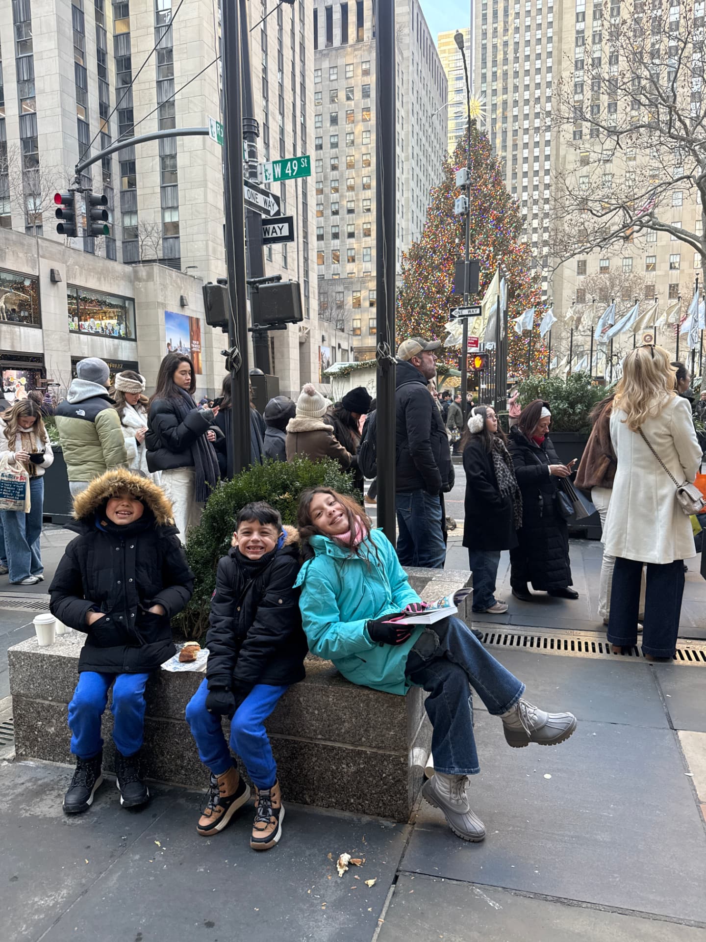The Hidalgo kids sitting on a bench near Rockefeller Center with the Christmas tree behind them during their Maman visit in New York City