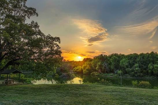 Sunset over the water at Trophy Club Park, with a covered picnic table, open grass, and trees along the shoreline